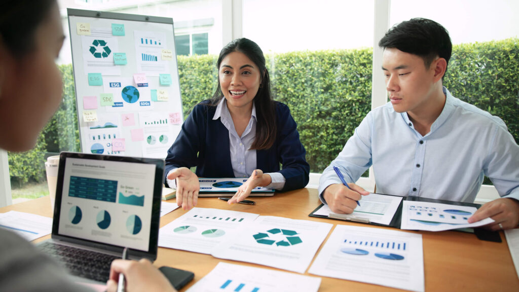 Reunião de profissionais analisando gráficos e dados de economia circular em papéis, quadro e tela de notebook.