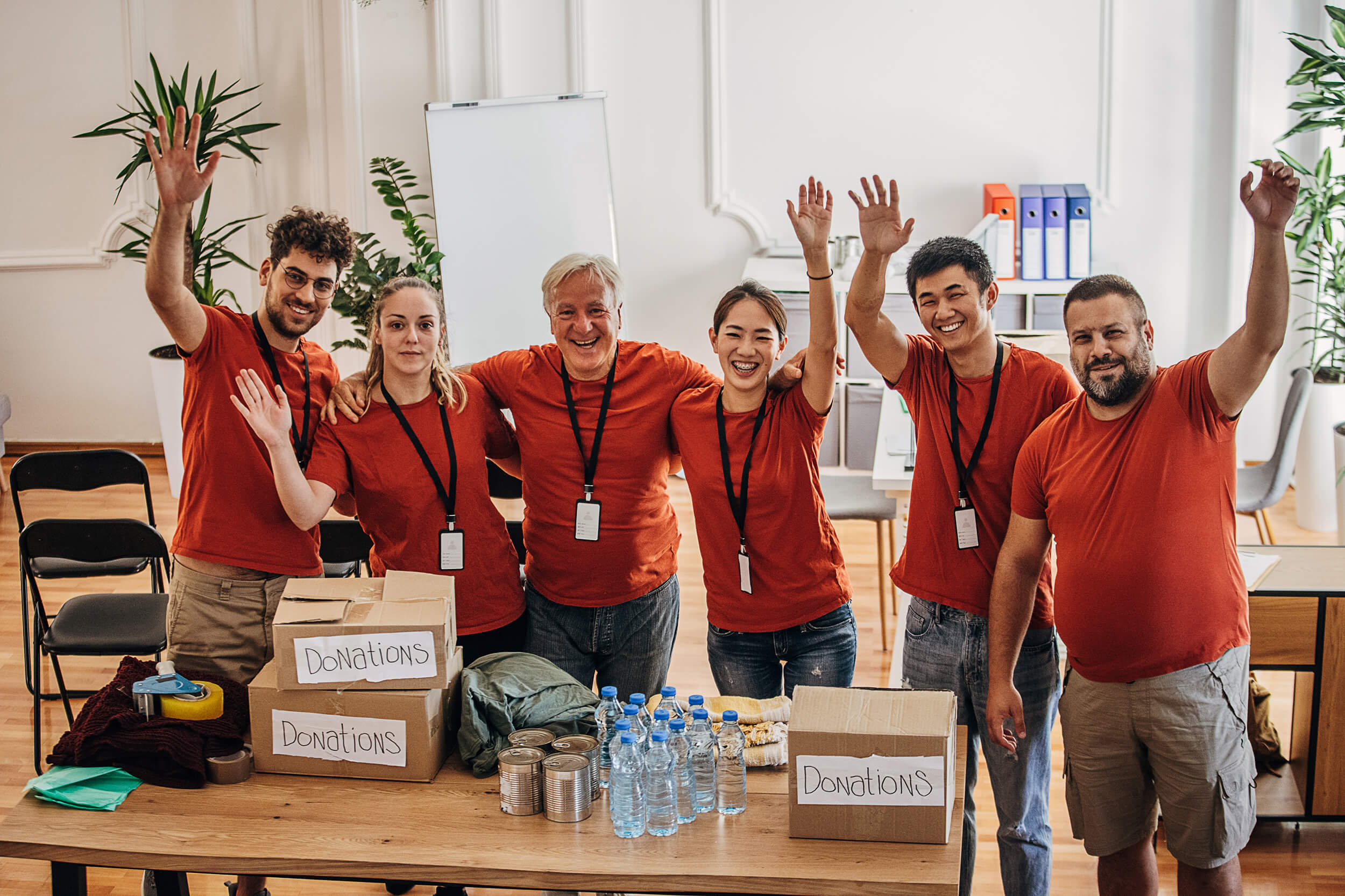 Voluntários de camiseta vermelha sorrindo em frente a uma mesa com 3 caixas de doações, além de garrafas de água e latas.
