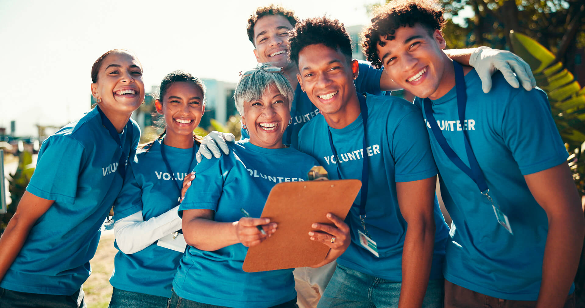 Grupo de voluntários em trabalho social usando camisetas azuis com a palavra “Volunteer”. Uma voluntária segura uma prancheta. Todos estão sorridentes.