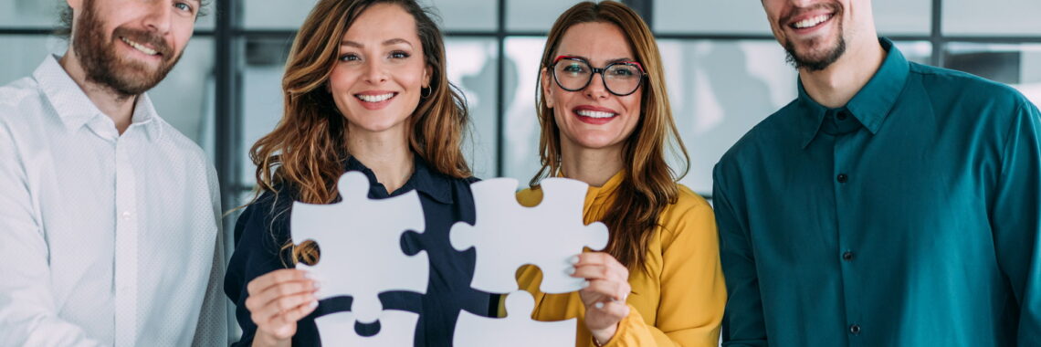 Quatro profissionais (dois homens e duas mulheres) vestindo roupa social. Todos estão sorridentes e segurando peças de quebra-cabeça, simbolizando parceria.