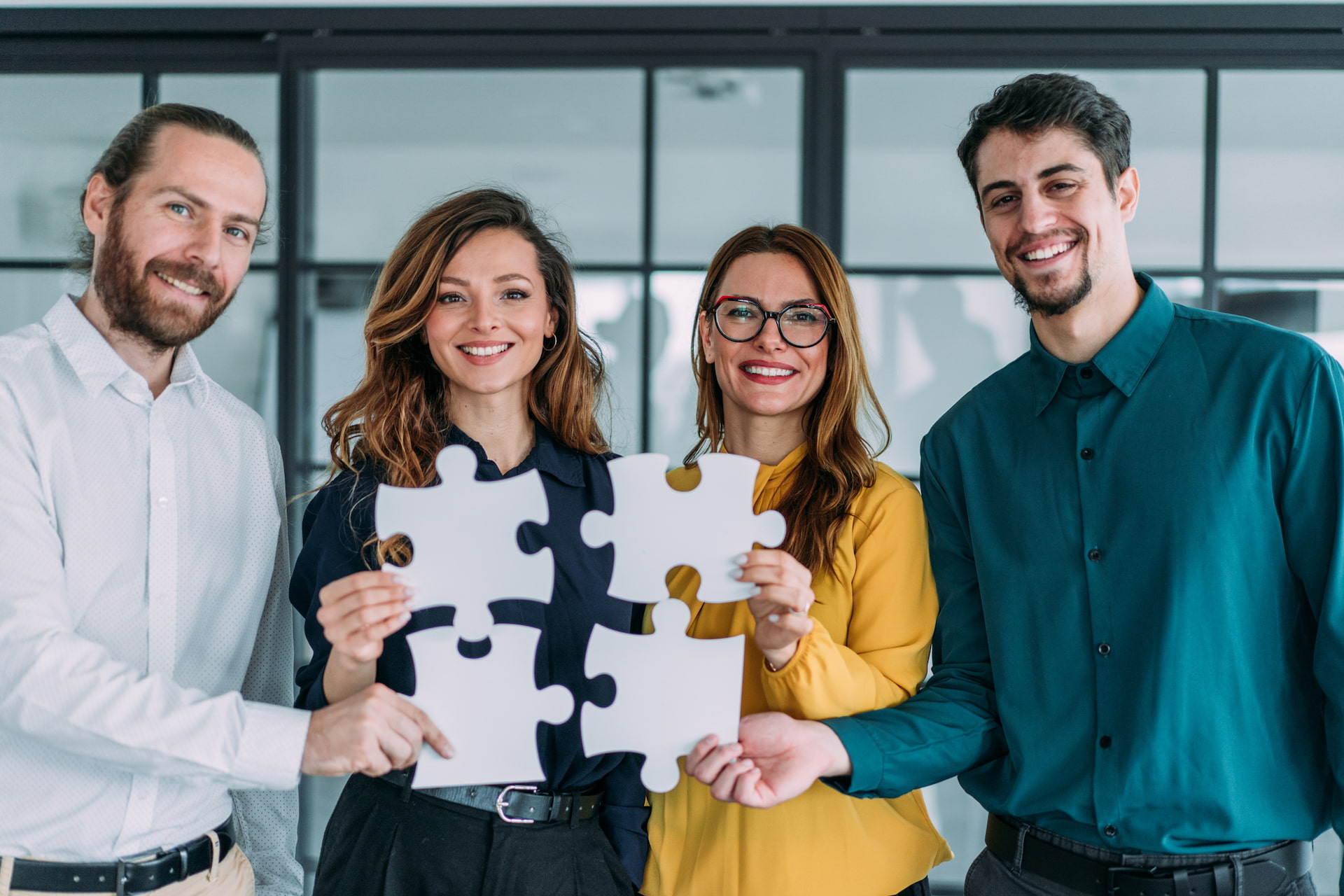 Quatro profissionais (dois homens e duas mulheres) vestindo roupa social. Todos estão sorridentes e segurando peças de quebra-cabeça, simbolizando parceria.