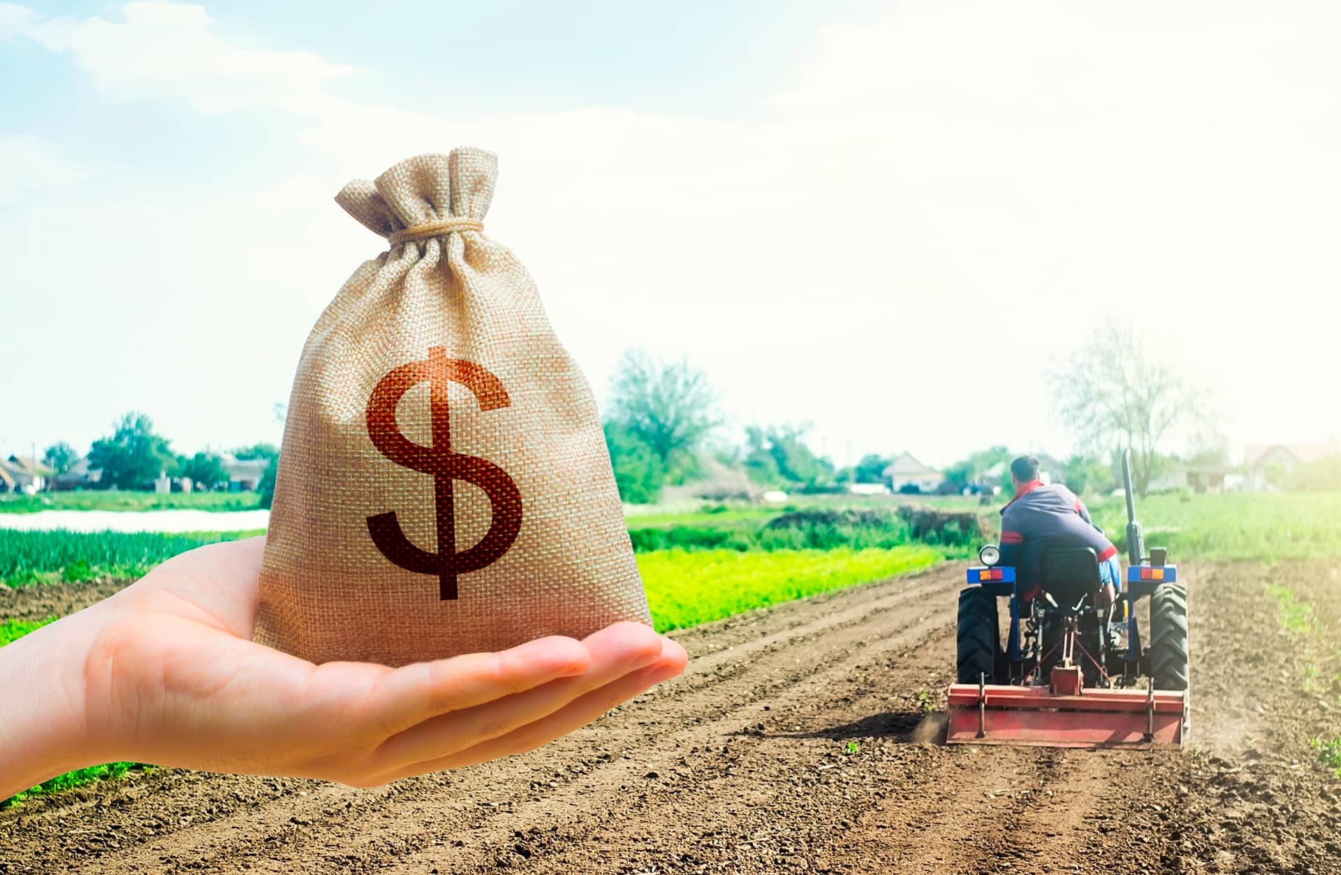 Mão segurando saco de dinheiro em lavoura com trator, representando financiamento no agronegócio.