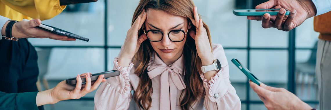 Executiva em ambiente de trabalho com sinais de estresse e sobrecarga emocional, cercada por colegas apontando celulares e canetas, ilustrando pressão psicológica.