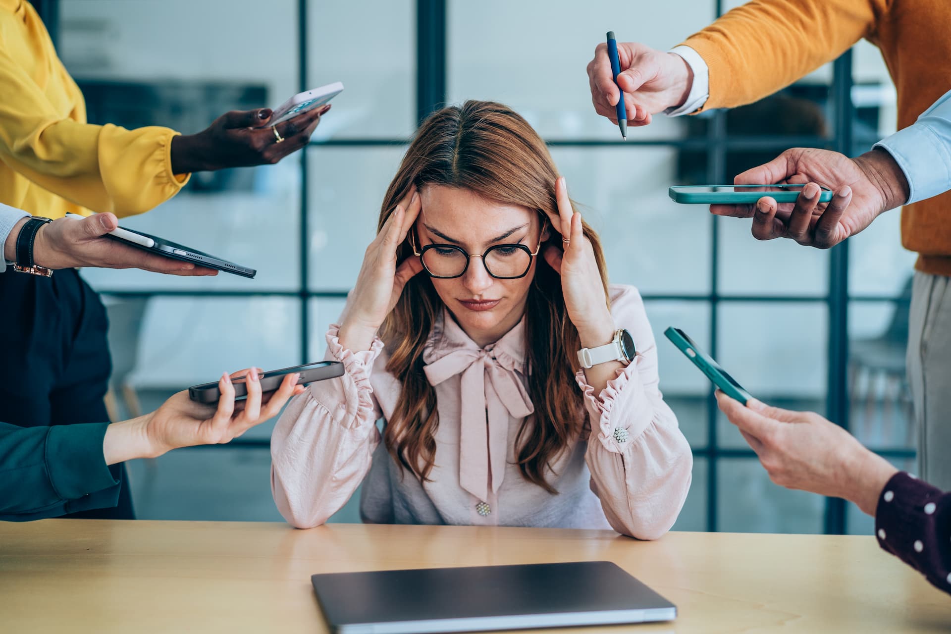 Executiva em ambiente de trabalho com sinais de estresse e sobrecarga emocional, cercada por colegas apontando celulares e canetas, ilustrando pressão psicológica.