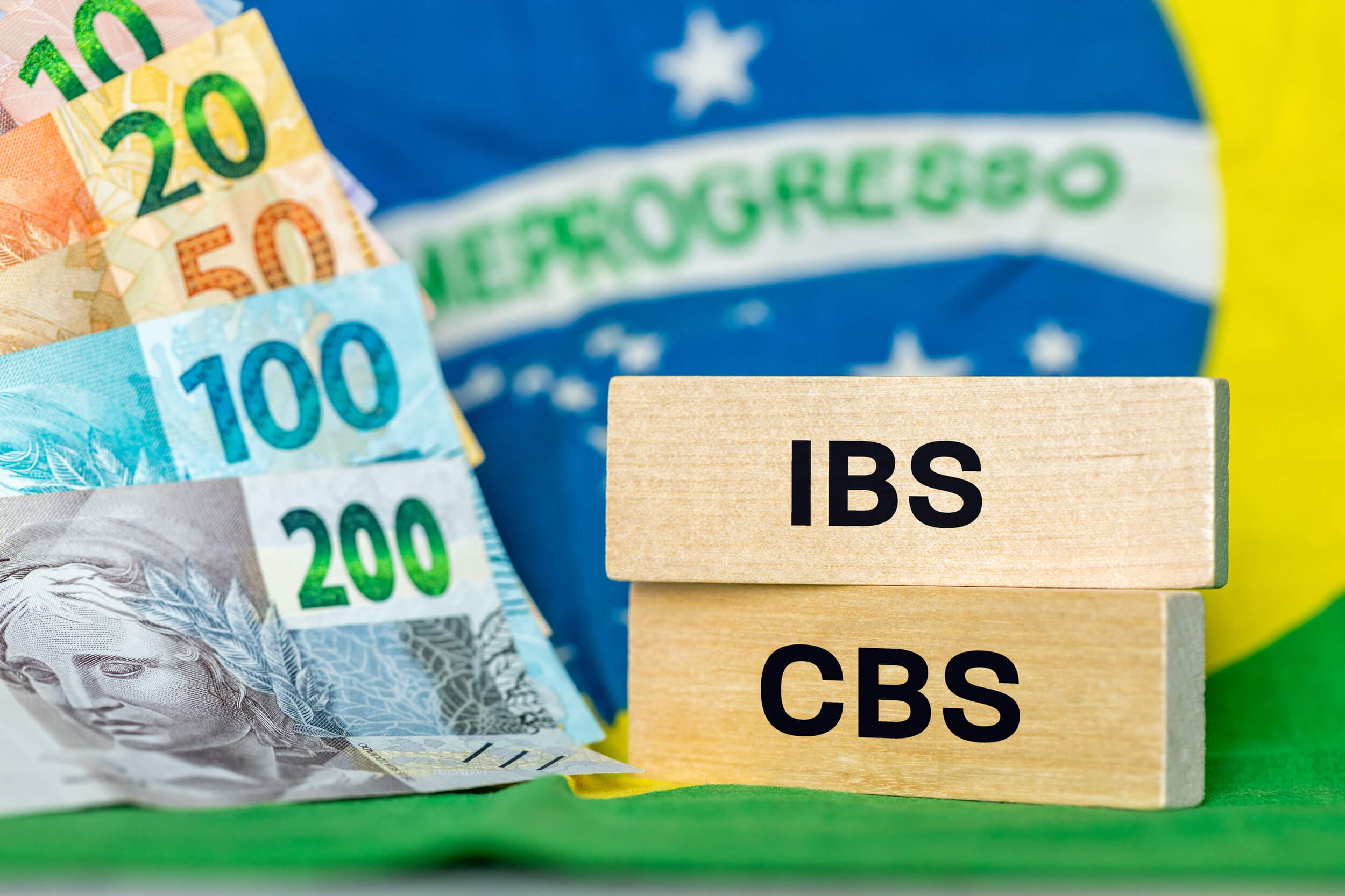 Cédulas de real e blocos de madeira com as siglas IBS e CBS sobre a bandeira brasileira.